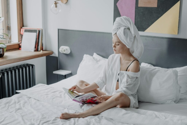 A young woman with washed hair wrapped in a towel, reading a magazine on a comfortable white bed.